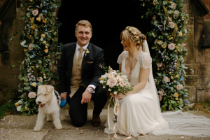 couple and dog pose in front of floral arch on wedding day